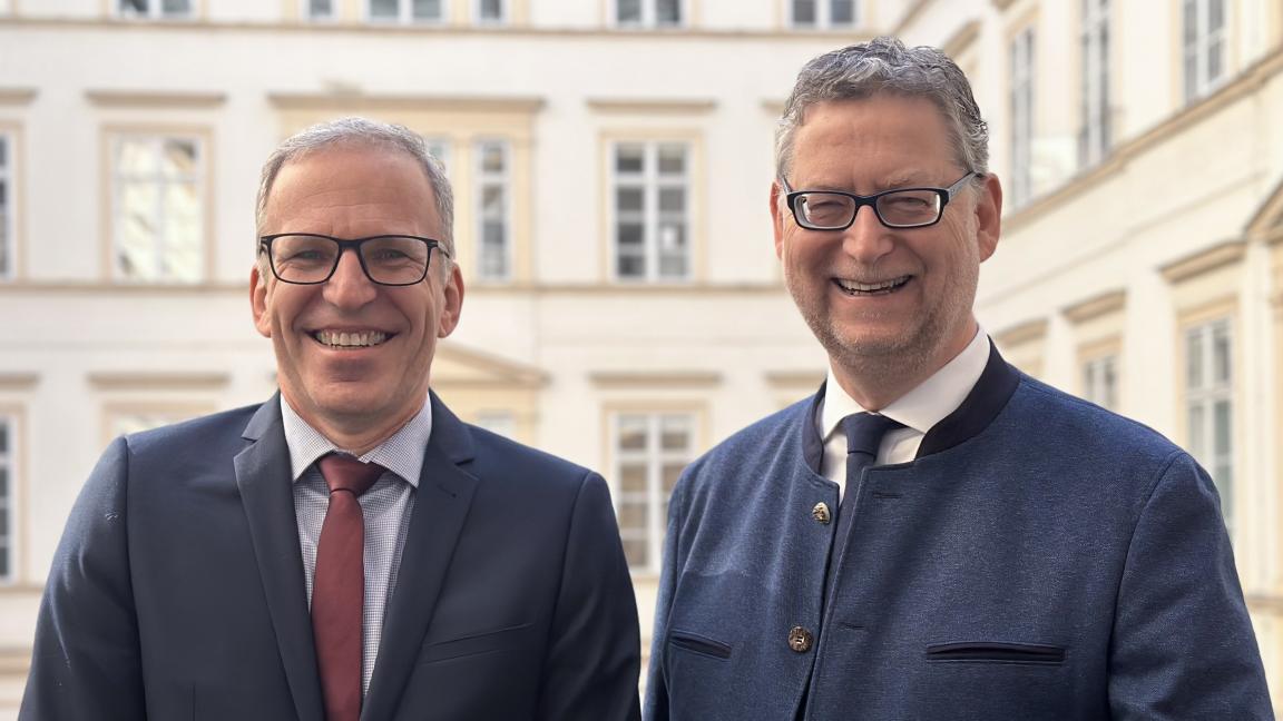 Two smiling men in suits stand in front of a light-colored building with many windows and a classical facade.