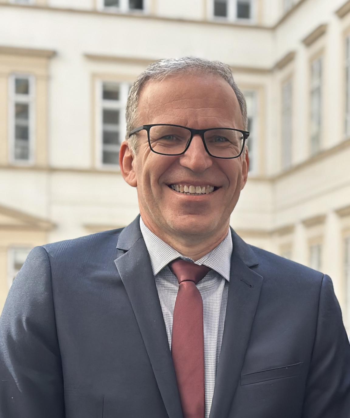 Portrait of a smiling man wearing glasses and a suit with a red tie in front of a historic building.