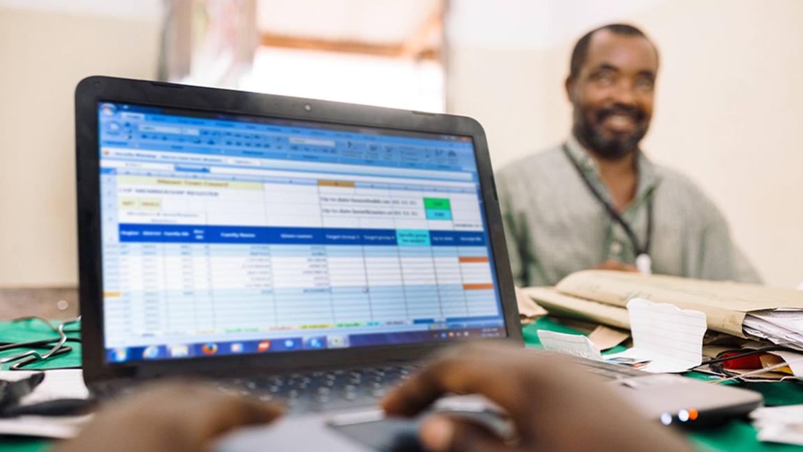 A person is typing on a laptop with an Excel spreadsheet, in the background a smiling man is sitting at a desk.
