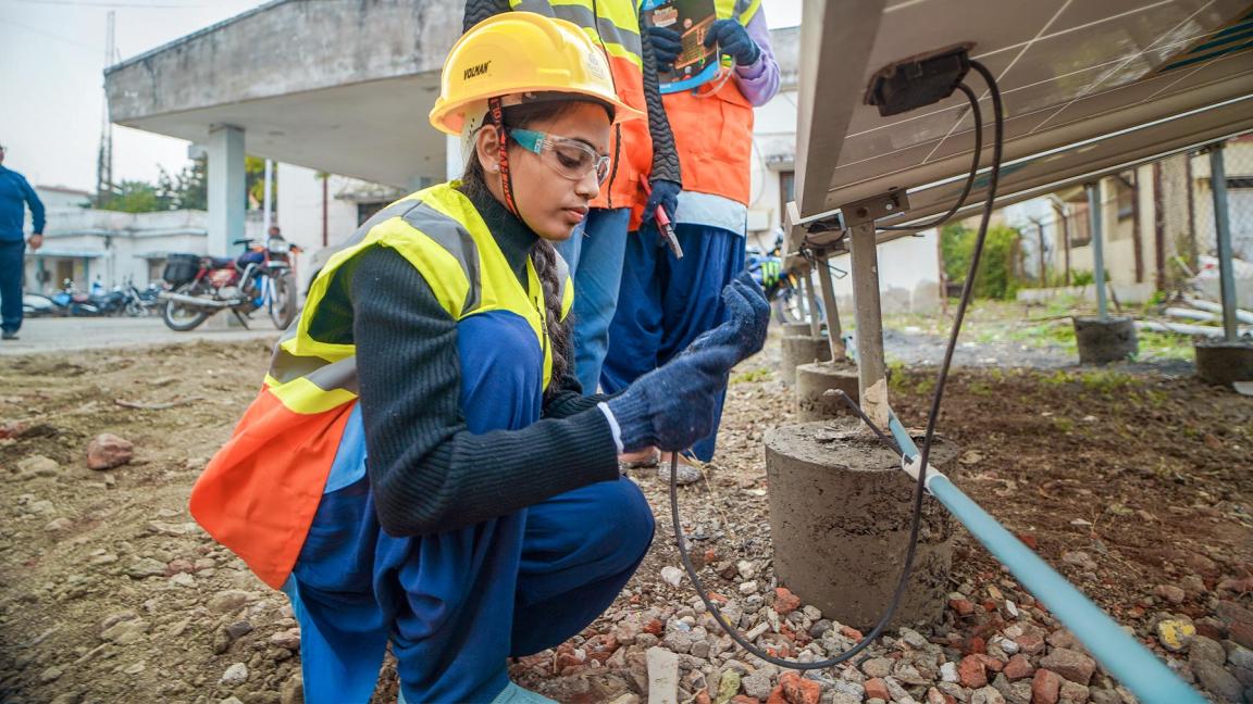 A young woman wearing protective clothing checks the cables under a solar panel; alongside her are two other people with tools and documents.