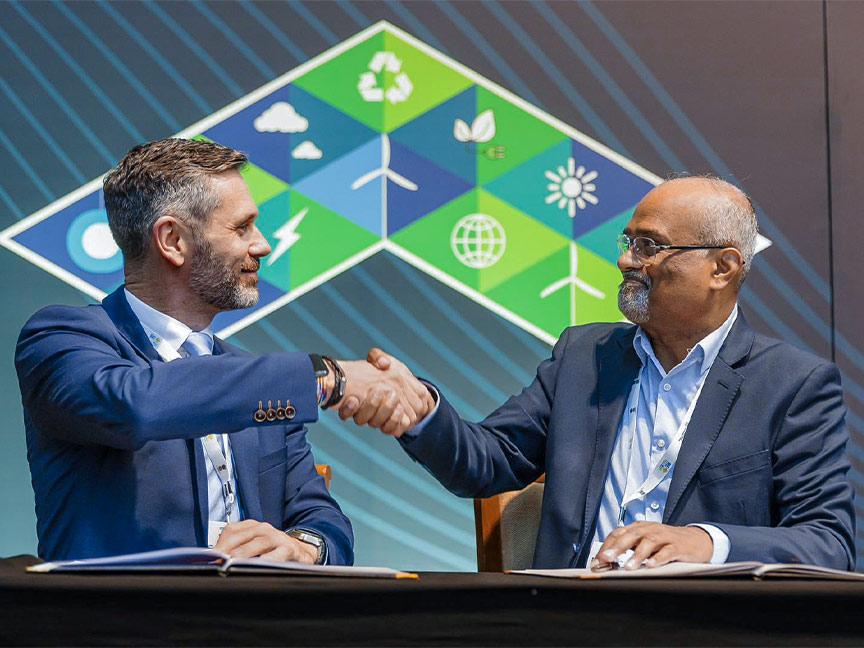 Two men in suits shake hands in agreement in front of a background of various symbols at the RE-INVEST 2024 conference.