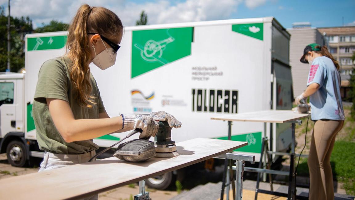 Two young women wearing protective masks use sanders to sand wooden boards in an outdoor setting in front of a mobile workshop vehicle featuring the logo ‘Tolocar’.