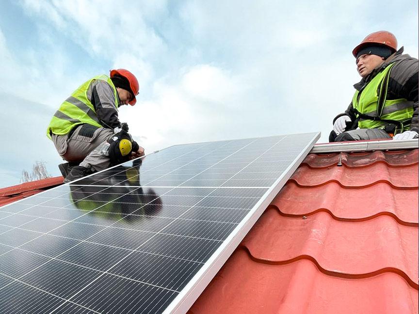 Two people in protective clothing and hard hats install a solar panel on a red tiled roof under a cloudy sky.