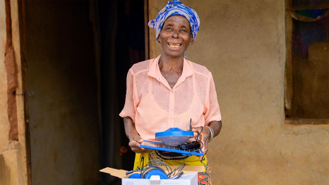 A smiling woman stands in front of a building holding a solar lamp set in her hand; in front of her is an opened package.