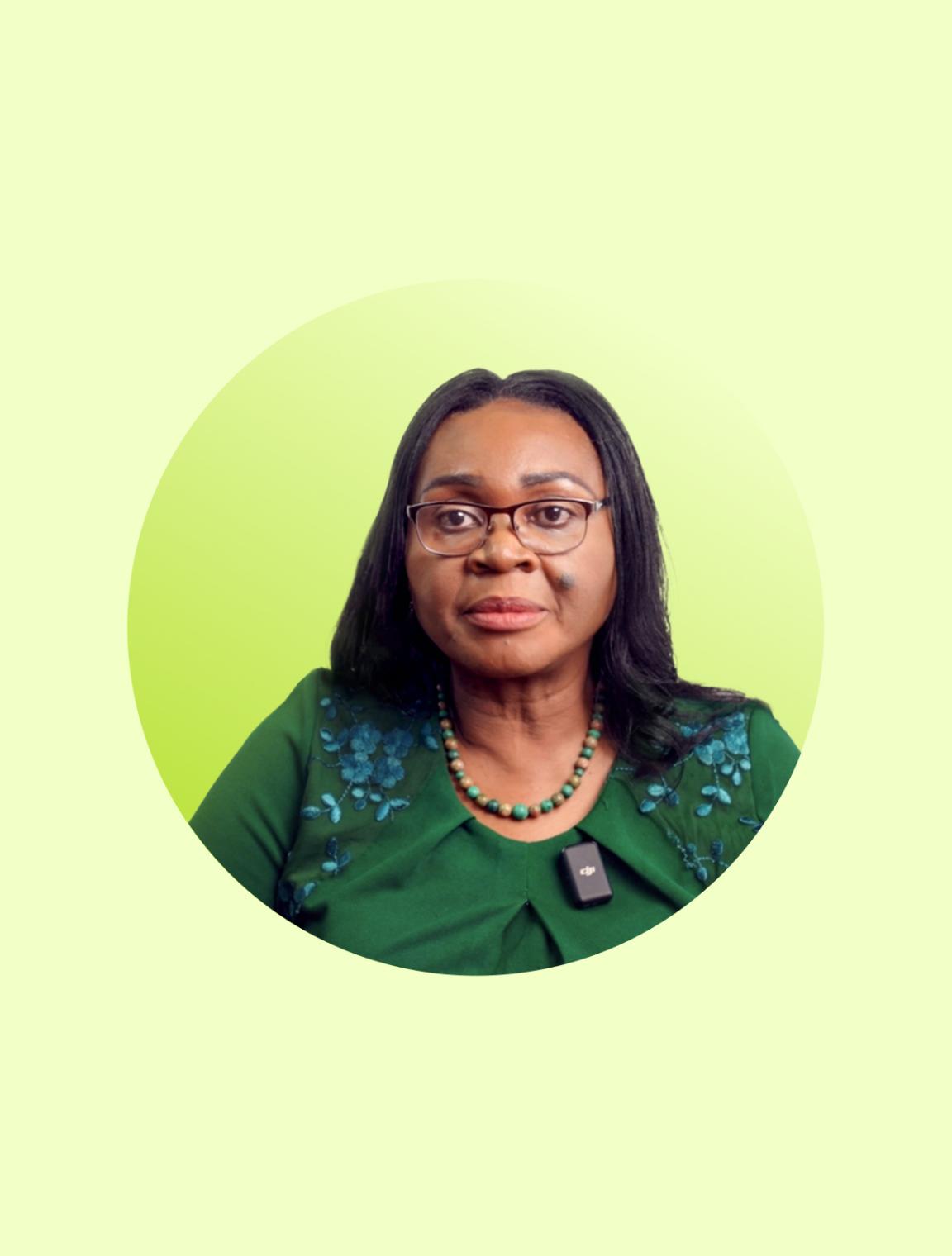 Porträt einer Frau mit Brille, grünem Kleid, Halskette und Ansteckmikrofon vor hellgrünem Hintergrund.