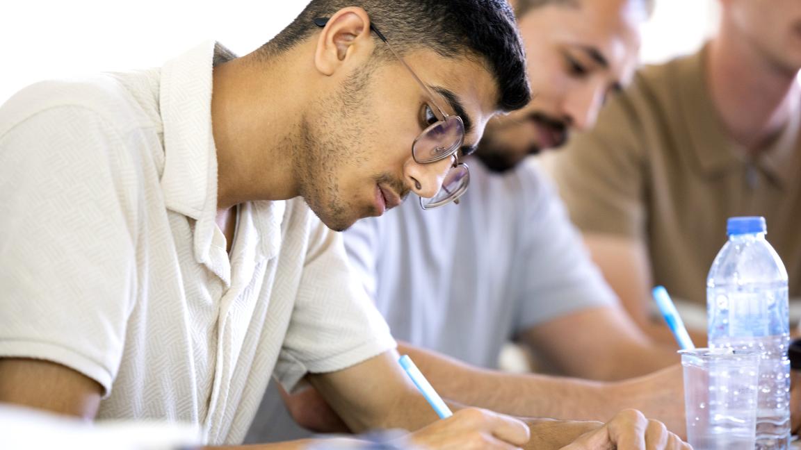 A young man with glasses writes attentively during a group activity, surrounded by other participants.