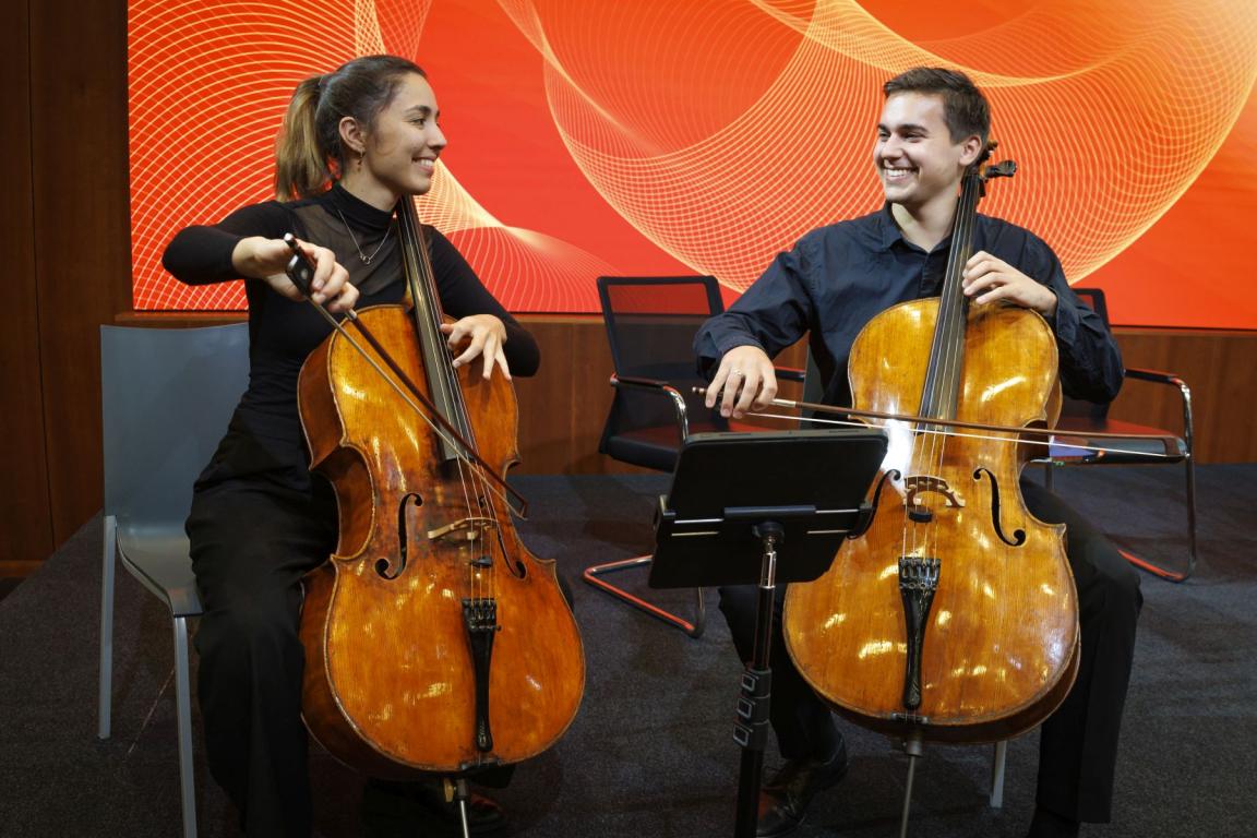 Two people playing the cello and smiling at each other