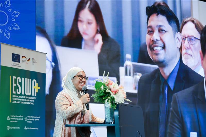 Eine Frau am Rednerpult neben einem Rollup mit der Aufschrift ESIWA+, im Hintergrund ein Foto mit Menschen