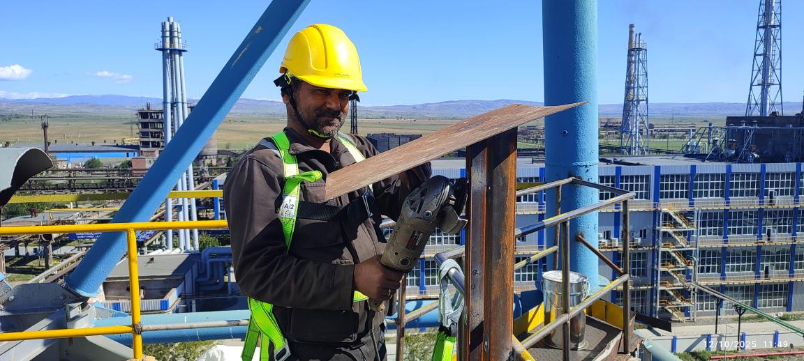 A man in a yellow helmet stands on a scaffold and grinds down a steel frame.