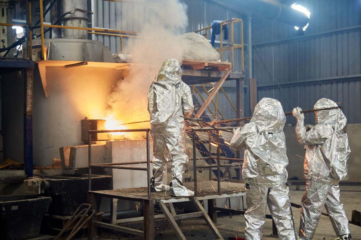 Three people in silver protective suits are working at a glowing furnace in a tin processing plant.
