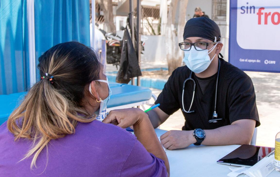 A doctor with a face mask, cap and stethoscope around his neck sits at a table opposite a woman
