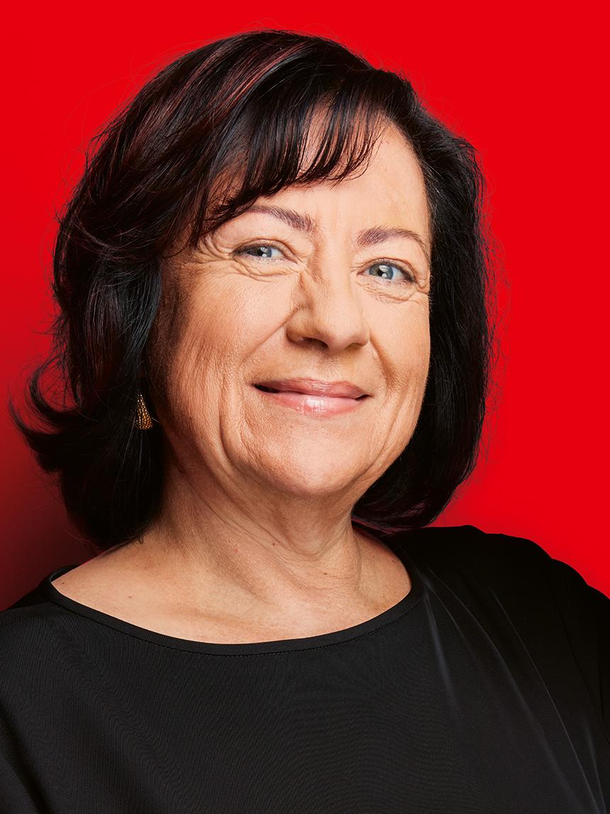 Portrait of a smiling older woman with dark shoulder-length hair and a black top in front of a bright red background.