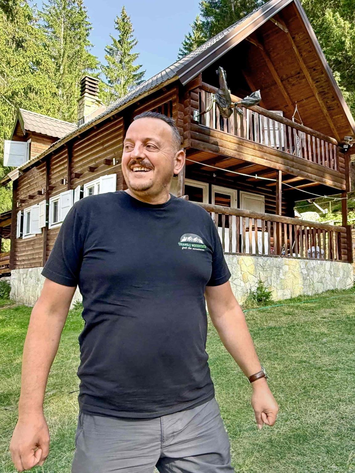 Ein lachender Mann steht vor einer rustikalen Holzhütte in einem Waldgebiet.