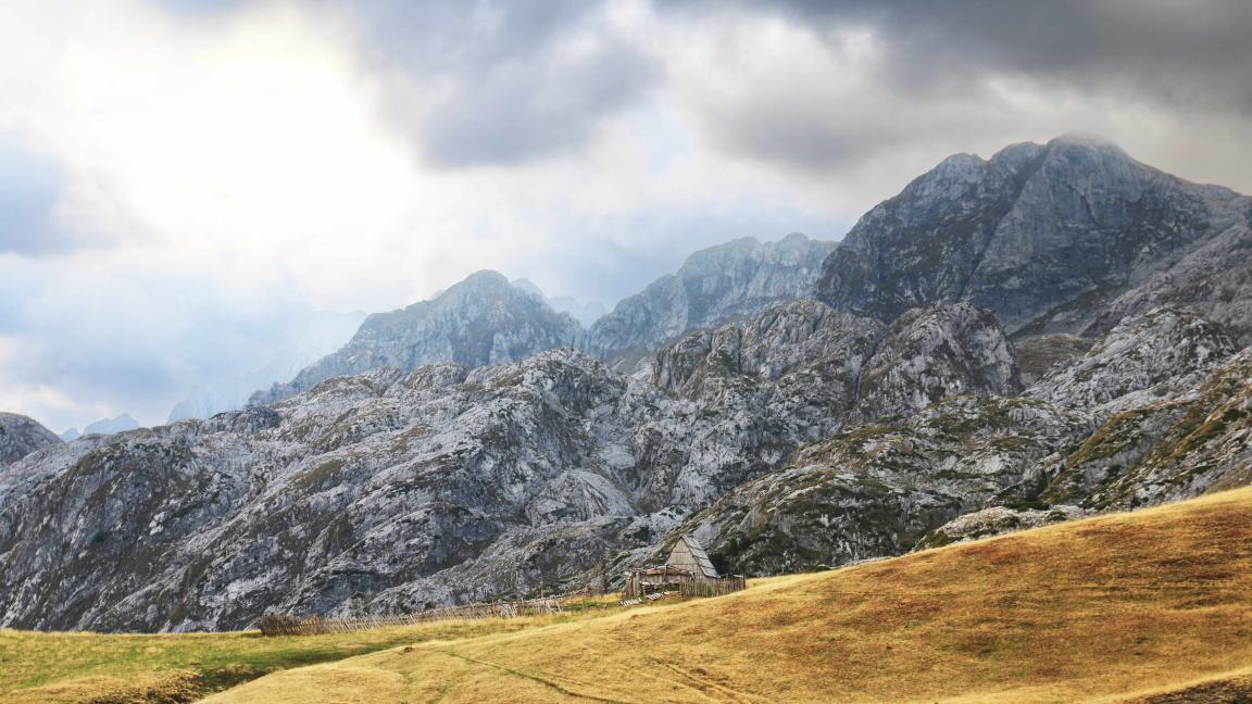 Eine kleine Holzhütte steht auf einer Wiese vor steilen, felsigen Berggipfeln unter dramatisch bewölktem Himmel.