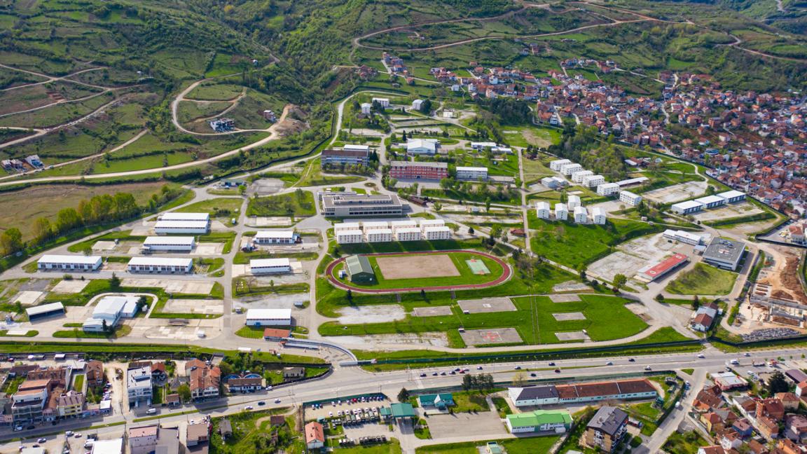 Luftaufnahme eines weitläufigen Geländes mit Gebäuden, Straßen und Sportplatz am Rand einer Stadt.