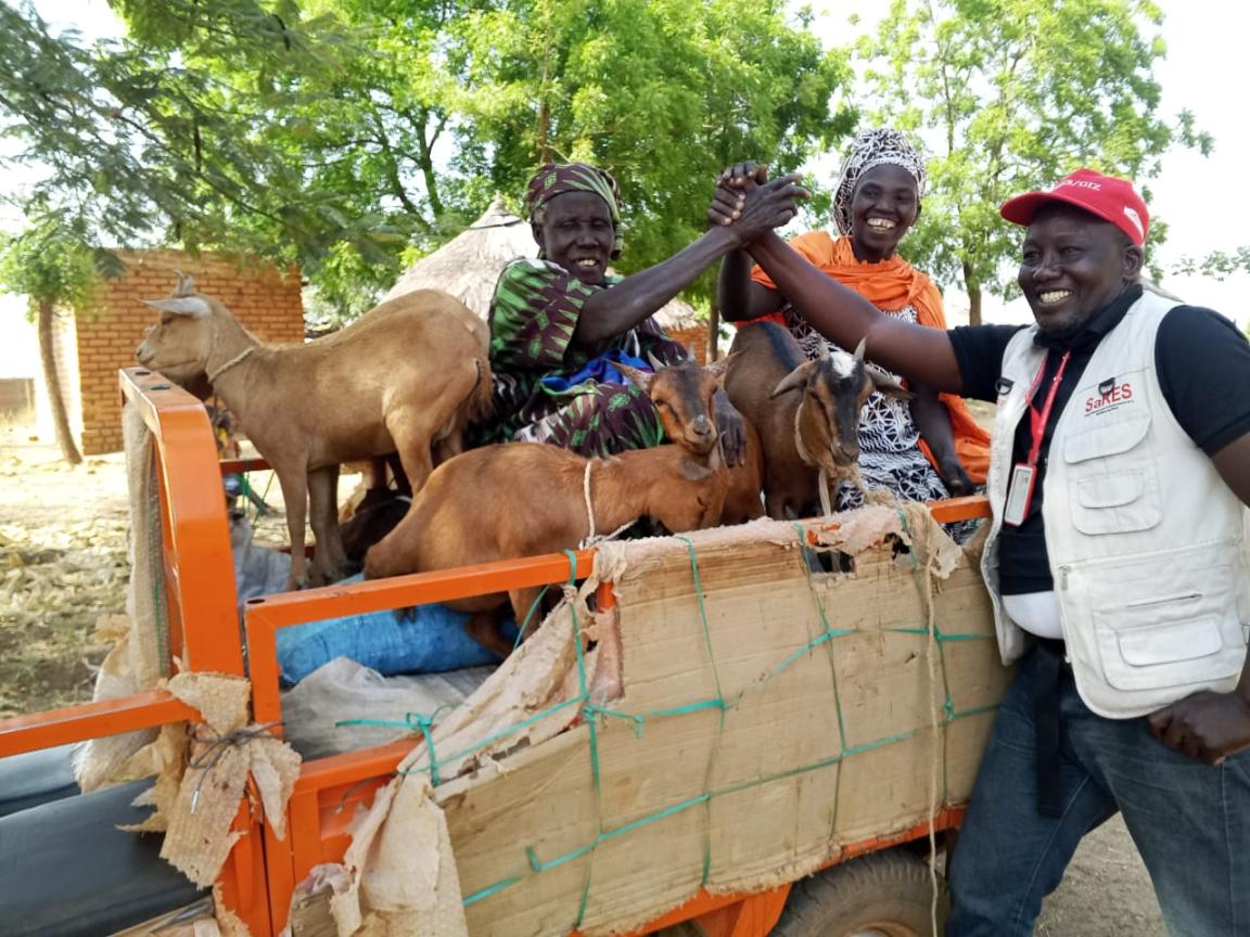 Drei lächelnde Menschen stehen an einem kleinen Karren, auf dem mehrere Ziegen transportiert werden. 