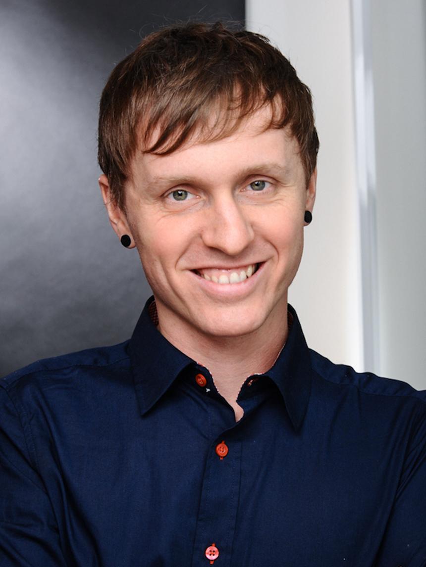 Portrait of a smiling man with short brown hair, ear studs, and a dark blue shirt in front of a neutral background.