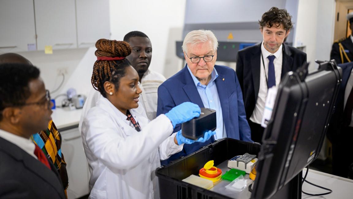 A group of people, including a prominent guest, watches a scientist demonstrate scientific equipment in the lab.