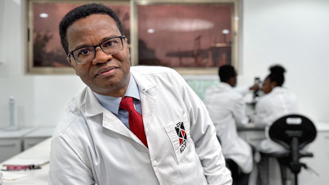 A man in a lab coat stands in the foreground with colleagues working behind him, showing a professional and focused atmosphere.