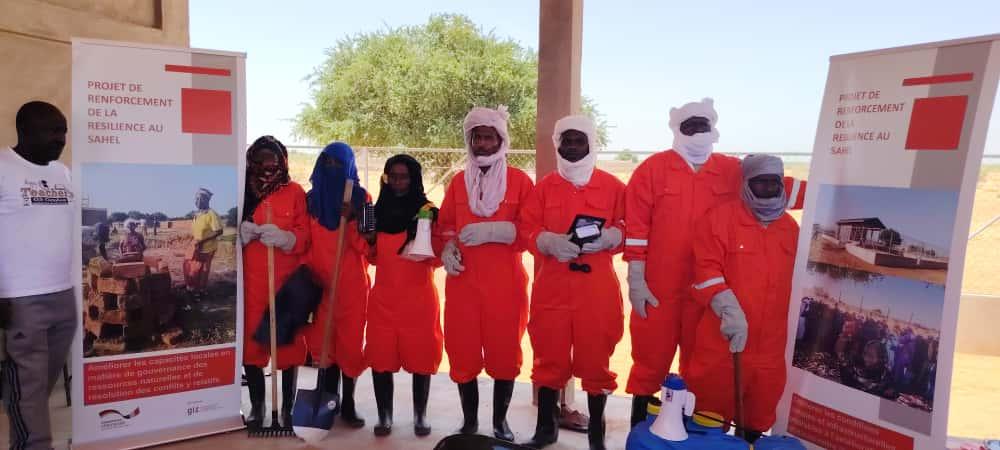 Eine Gruppe von sieben Personen in orangen Schutzanzügen steht zwischen zwei Informationsbannern im Freien. 