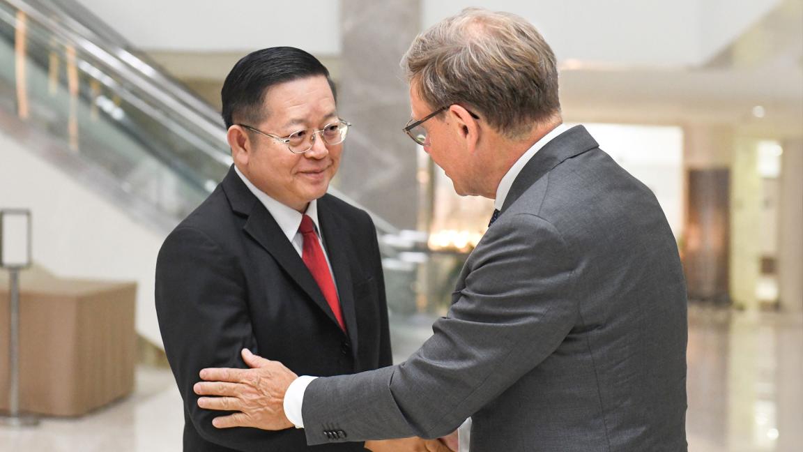 Zwei Männer in Anzügen begrüßen sich in einem modernen Gebäude mit Rolltreppe im Hintergrund bei einem formellen Treffen.