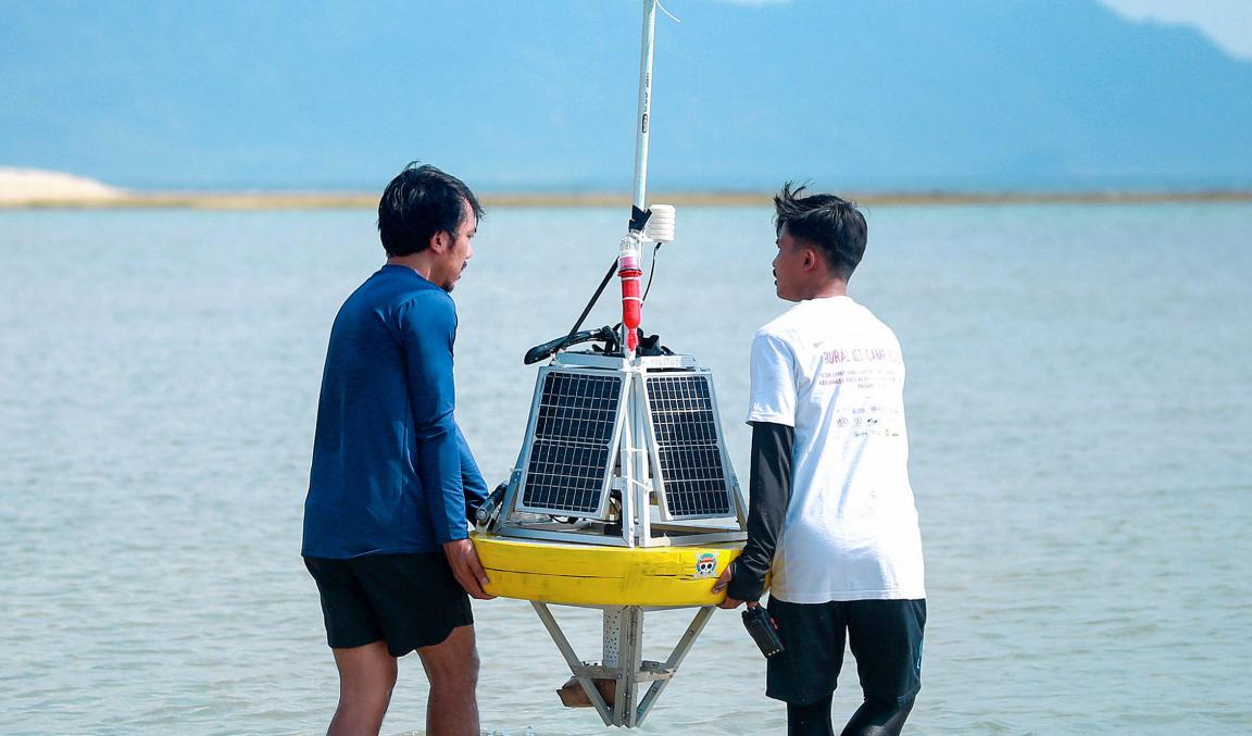 Zwei Männer tragen gemeinsam ein schwimmendes Messgerät mit Solarpanel auf einem Gewässer bei klarem Himmel.