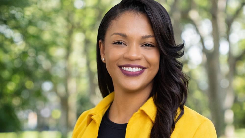 Portrait of a smiling woman with long dark hair wearing a yellow blazer and black top in front of a blurred green park background in daylight.