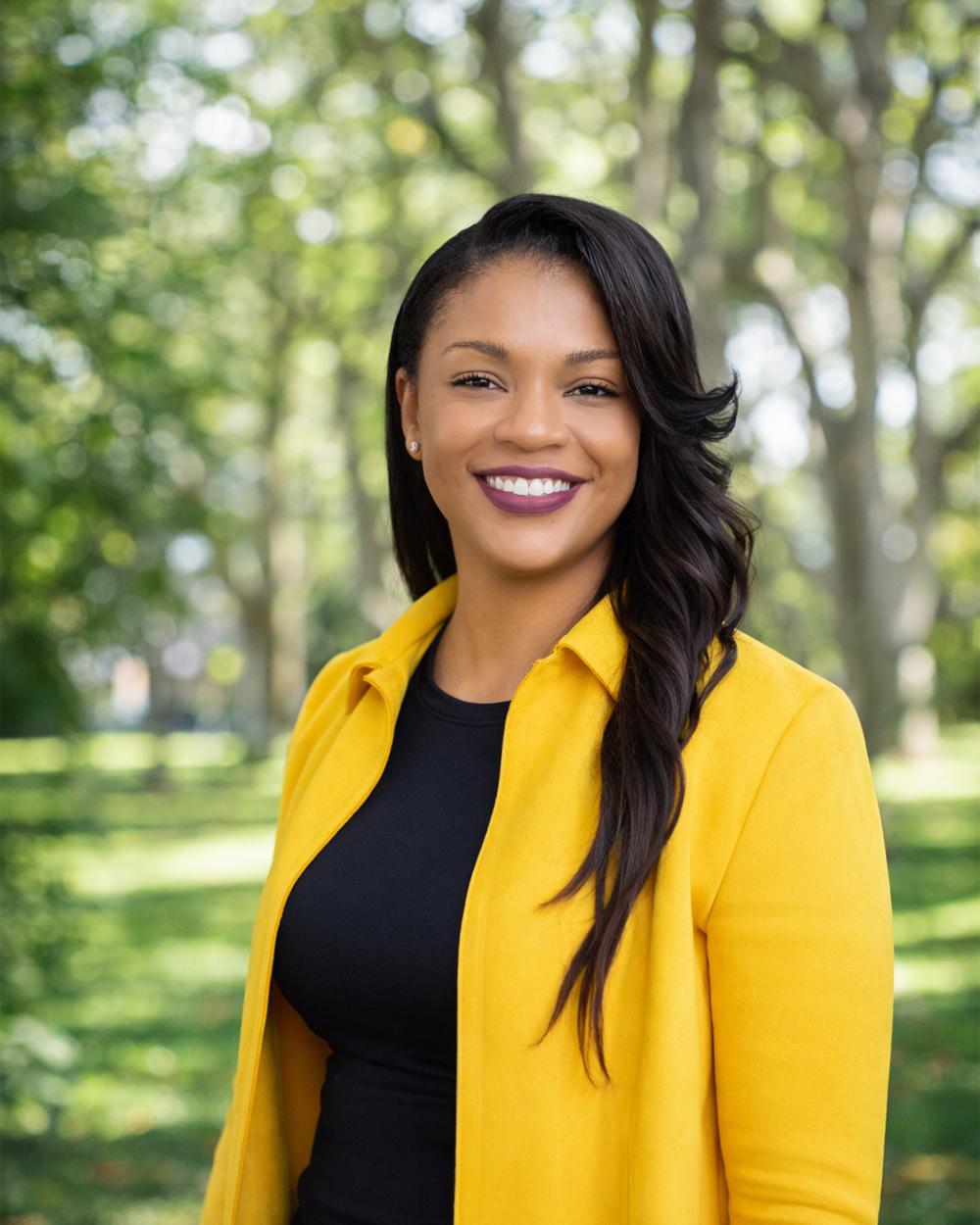 Portrait of a smiling woman with long dark hair wearing a yellow blazer and black top in front of a blurred green park background in daylight.