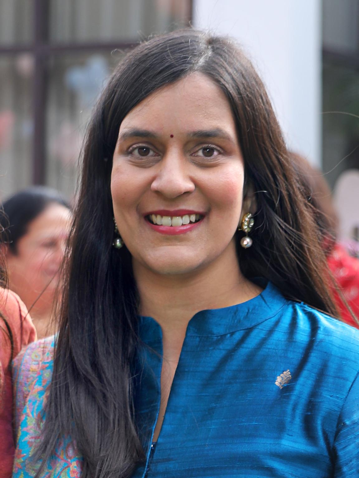 Eine lächelnde Frau mit langen dunklen Haaren und blauem Oberteil blickt frontal in die Kamera, während sich Menschen unscharf im Hintergrund befinden.