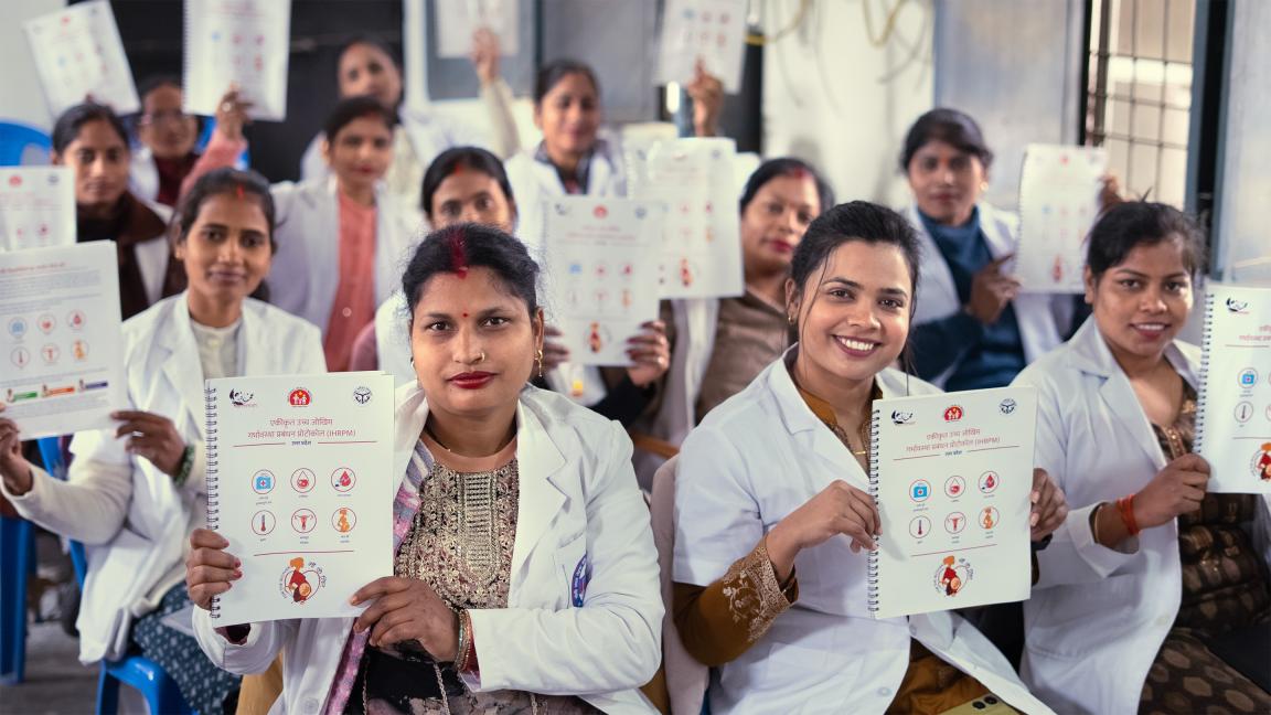 Eine Gruppe von Frauen in weißen Kitteln sitzt in einem Schulungsraum und hält Hefte mit Gesundheitsgrafiken in die Kamera.