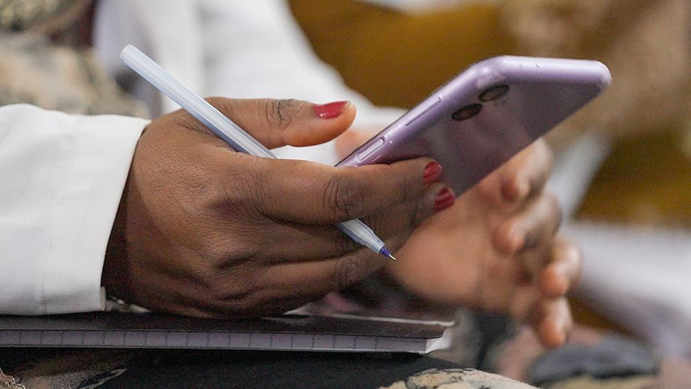 Close-up of a hand holding a purple smartphone while the other hand holds a pen. A notebook is visible in the background, with the focus on the hands.