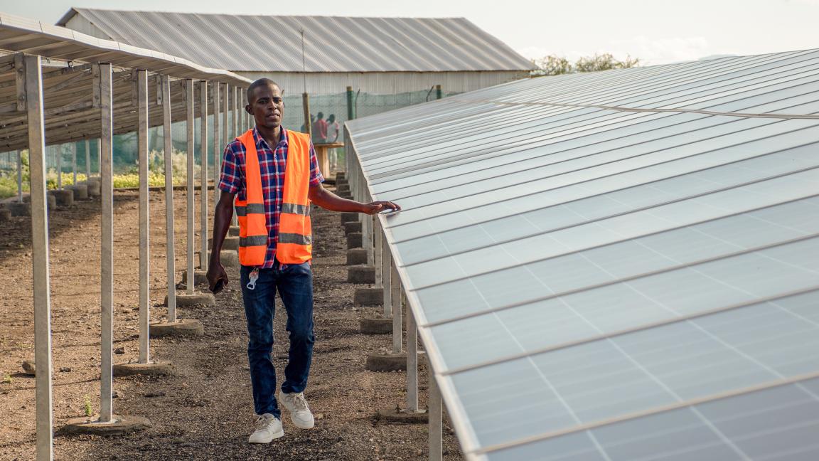 Man wearing an orange safety vest and checkered shirt standing next to a row of solar panels at an outdoor site with metal structures and buildings in the background.