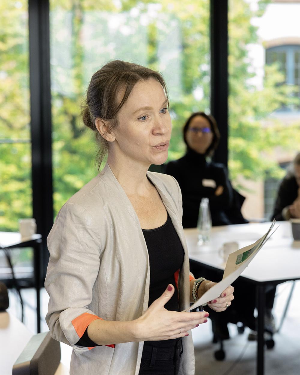 Frau mit zusammengebundenen Haaren hält ein Dokument in der Hand und spricht in einem hellen Seminarraum mit großen Fenstern, während im Hintergrund weitere Personen sitzen.