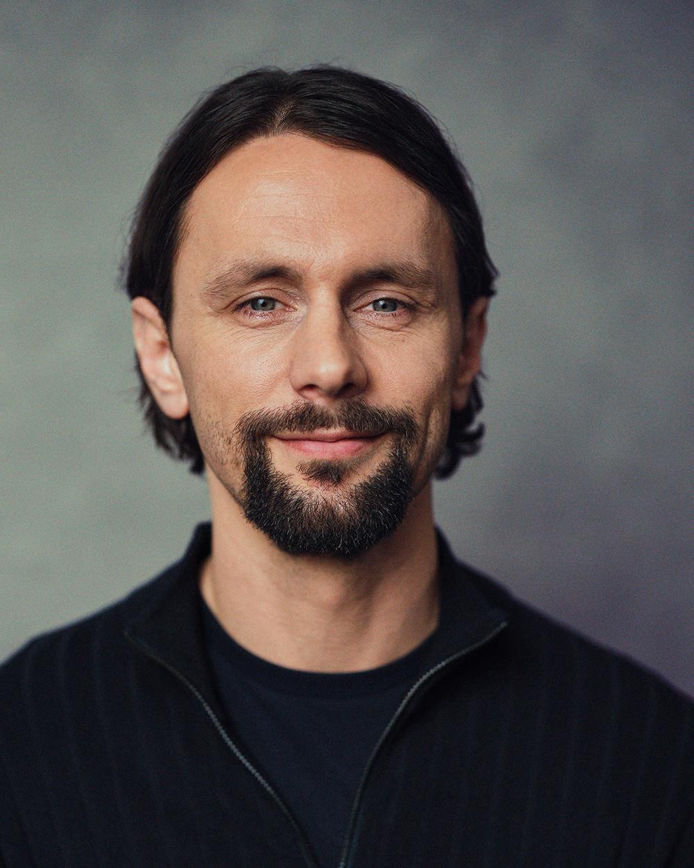 Porträtaufnahme eines Mannes mit halblangem dunklem Haar und Bart, der ein dunkelblaues Oberteil mit Reißverschluss trägt und in die Kamera lächelt.