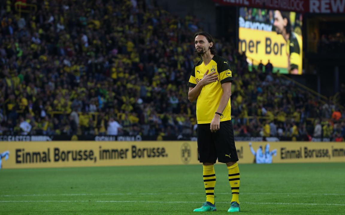 Ein Fußballspieler im gelb-schwarzen Trikot von Borussia Dortmund steht auf dem Rasen, hält sich die Hand ans Herz und blickt emotional in Richtung der Zuschauertribüne mit dem Banner „Einmal Borusse, immer Borusse.“