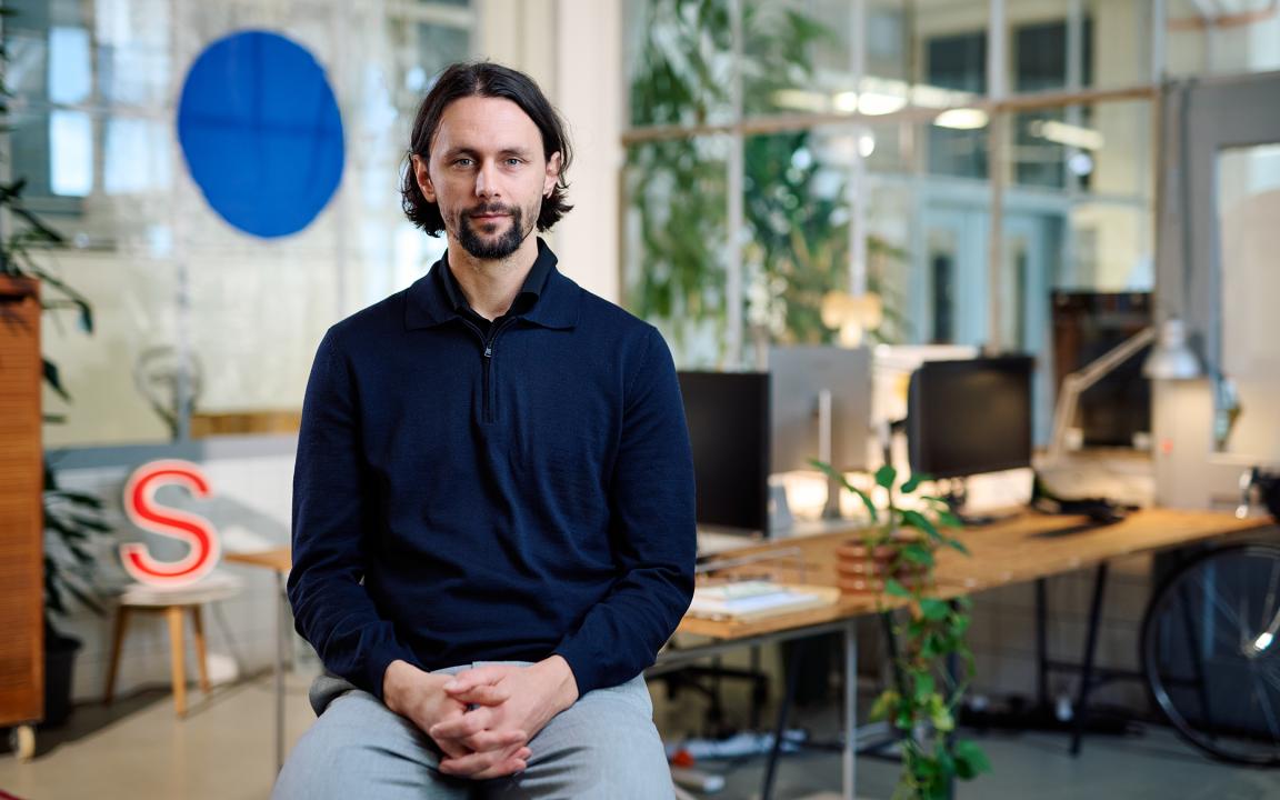 Ein Mann in einem dunkelblauen Pullover mit Reißverschluss sitzt mit gefalteten Händen in einem modernen, begrünten Büro und blickt ruhig in die Kamera.