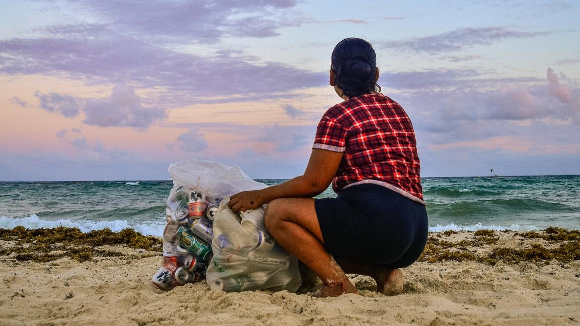 Eine Frau hockt bei Sonnenuntergang am Strand neben einem mit gesammelten Dosen und Plastikflaschen gefüllten Sack und blickt aufs Meer.