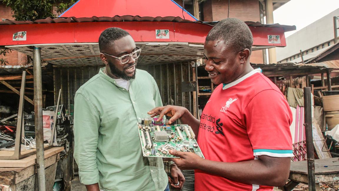 Zwei Männer betrachten lächelnd eine Computer-Platine auf einem Elektroschrott-Markt.