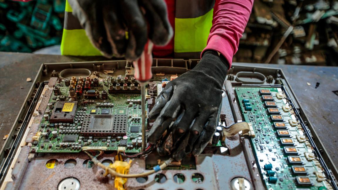 Nahaufnahme von behandschuhten Händen, die mit einem Schraubendreher eine elektronische Platine aus einem Altgerät demontieren.