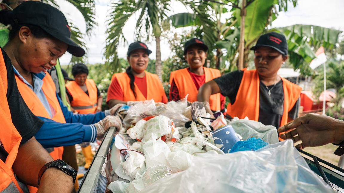Eine Gruppe von Frauen in orangefarbenen Warnwesten sortiert gemeinsam Plastikmüll an einem Sammelpunkt im Freien.