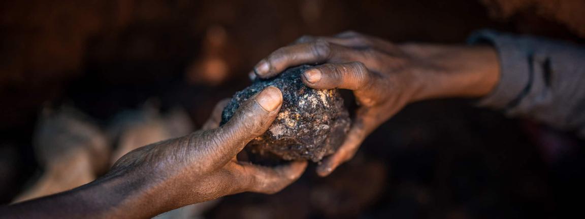 Zwei Hände halten einen dunklen, unbearbeiteten Gesteinsbrocken in einer schwach beleuchteten Umgebung.