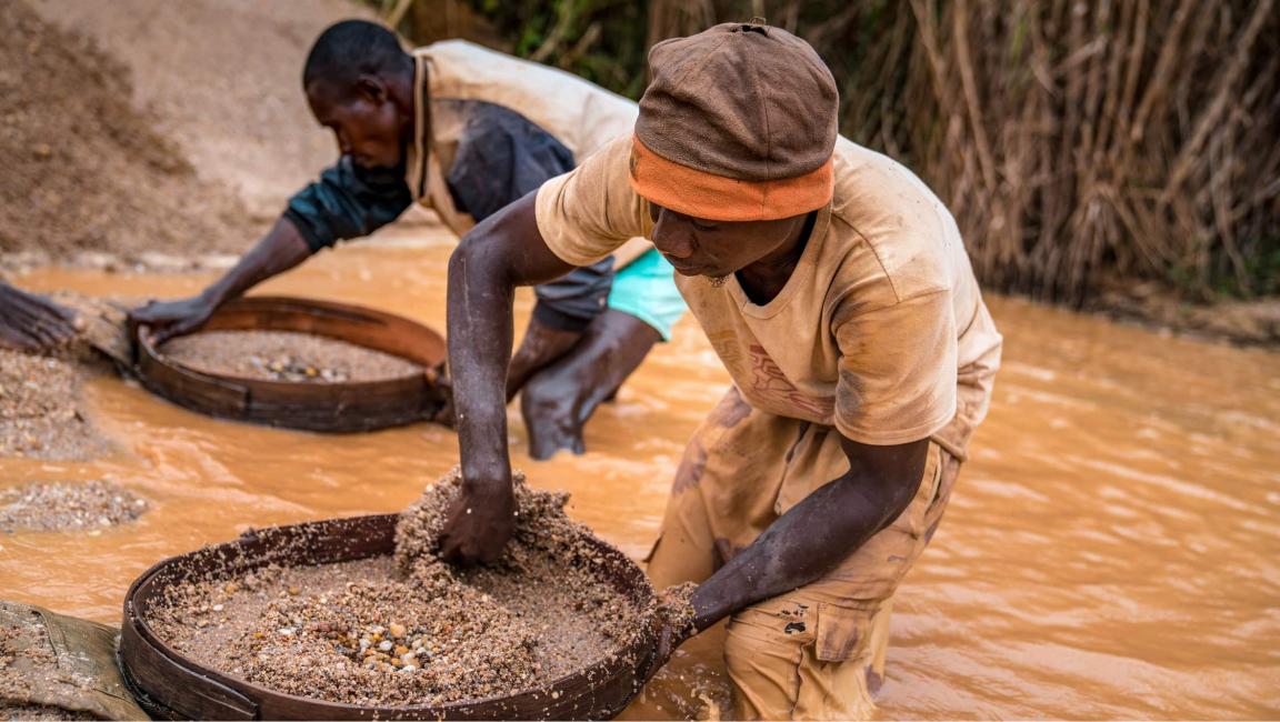 Zwei Arbeiter sieben mit großen Schalen Rohstoffe in schlammigem, orangefarbenem Wasser im handwerklichen Bergbau.