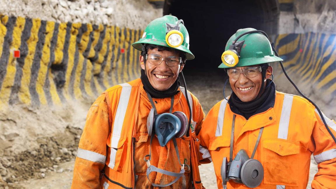 Zwei lächelnde Bergarbeiter in orangefarbener Schutzkleidung mit grünen Helmen und Stirnlampen stehen vor einem Stolleneingang.