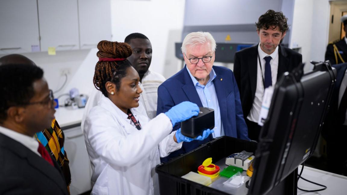 Eine Gruppe von Personen, darunter ein prominenter Gast, beobachtet eine Wissenschaftlerin, die ein wissenschaftliches Gerät im Labor vorführt.