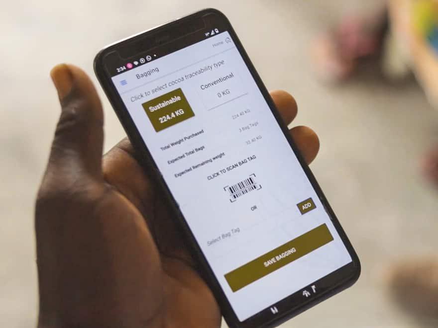 Eine Hand hält ein Smartphone, das eine Kakao-Rückverfolgbarkeits-App mit Barcode-Scanner und der Anzeige „224 KG Sustainable“ zeigt.