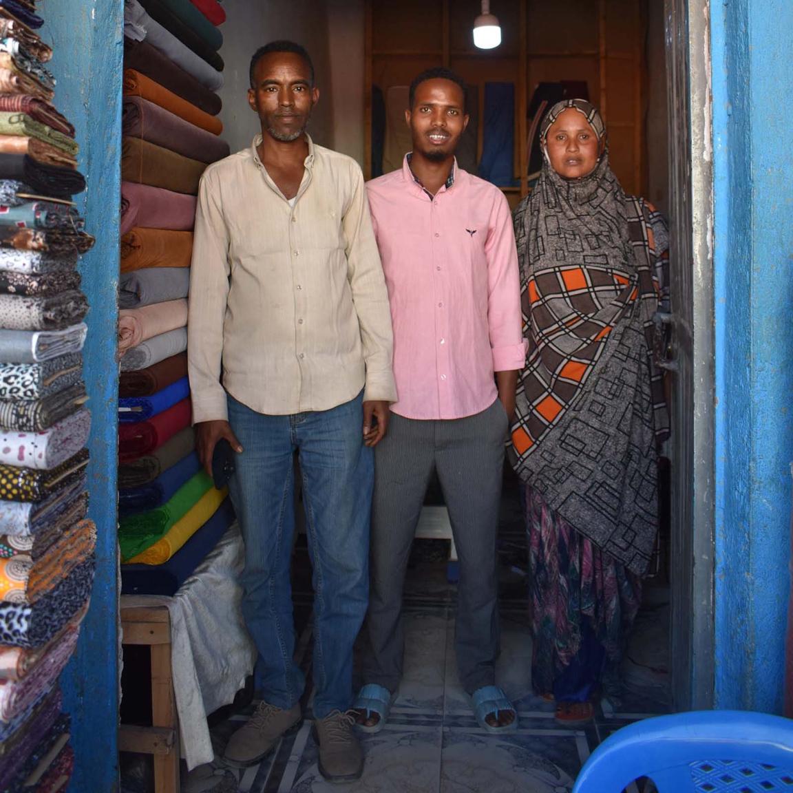 Two men and a woman are standing in a shop. To their right, piles of colourful fabric are stacked high.