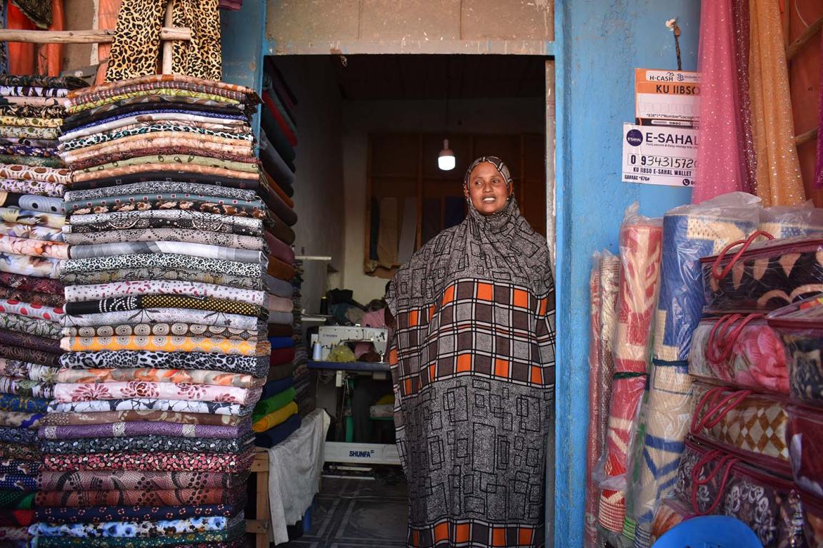 A woman in a colourful dress is standing next to a pile of fabrics in a small shop.