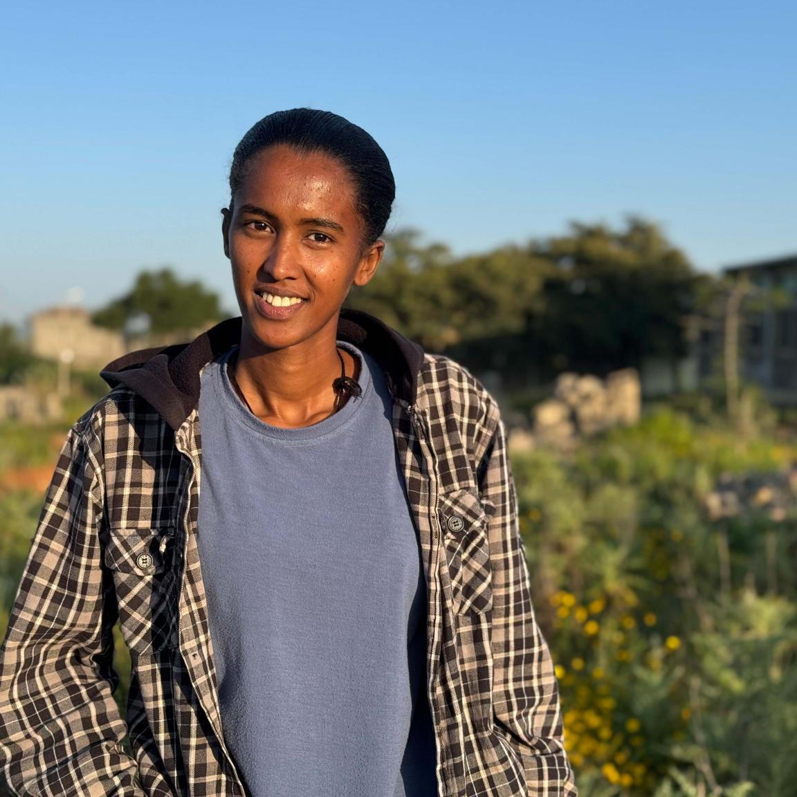 A young woman is standing in a green, blossoming landscape. She is smiling at the camera.