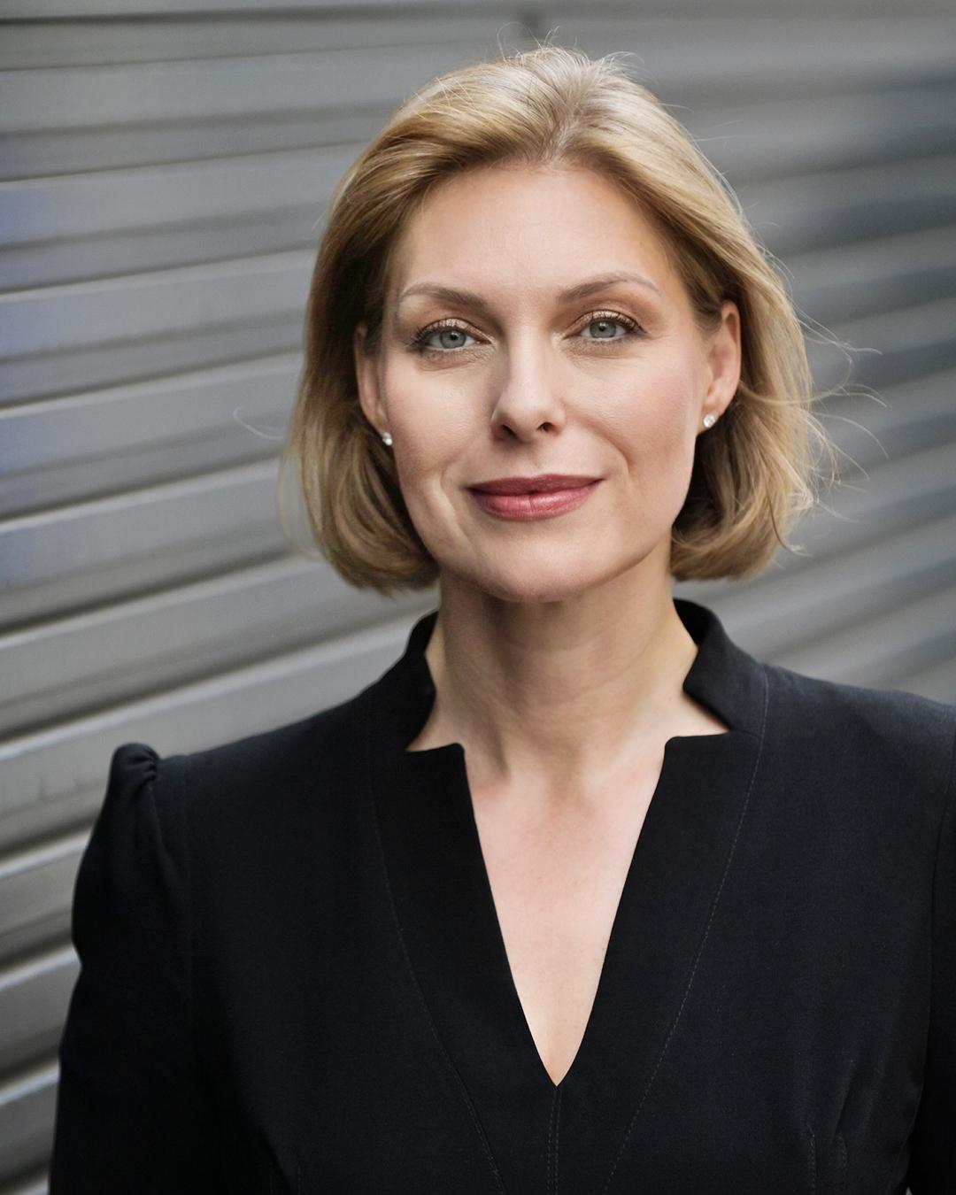 Porträt einer Frau mit kurzem blondem Bob in schwarzer Bluse, die leicht lächelt, vor einem grauen, unscharfen Hintergrund mit horizontalen Linien.