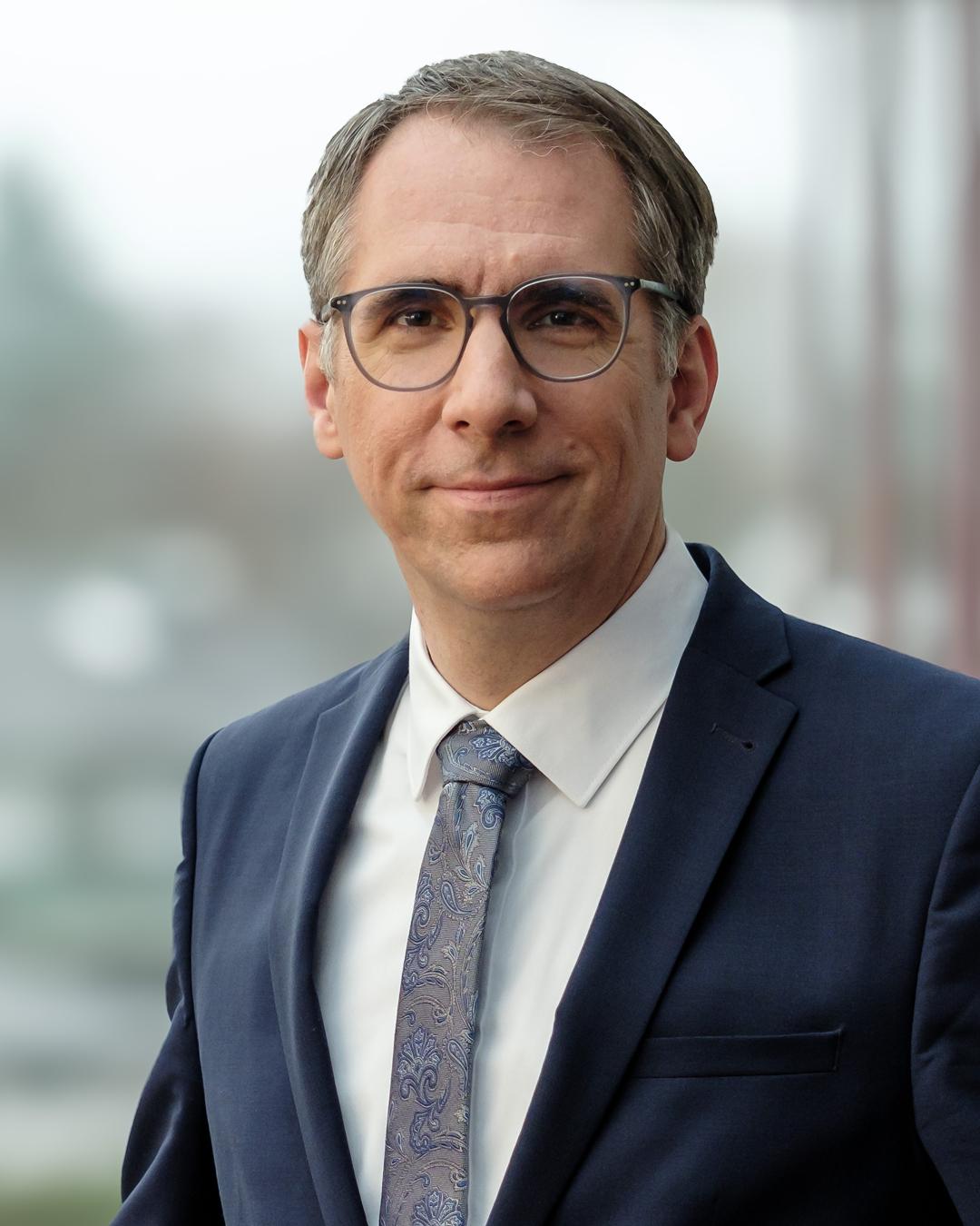 Porträt eines Mannes mit kurzer grauer Frisur und Brille im dunkelblauen Anzug, weißem Hemd und gemusterter Krawatte vor hellem, unscharfem Hintergrund.